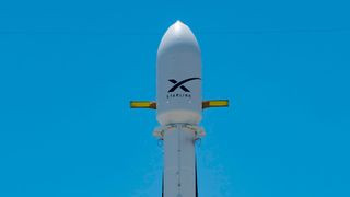 A white rocket topped with a payload fairing stands on the launch pad in a close-up set against a blue sky.